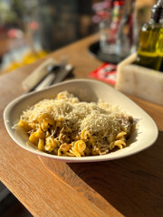 Bowl of pasta topped generously with grated cheese under warm sunlight. Rustic wooden table adds to the inviting and homely atmosphere