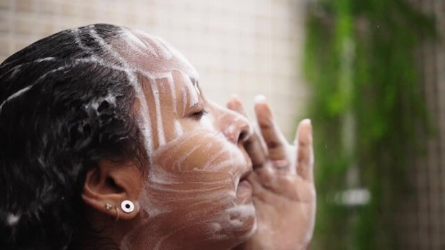 Close up of a young black woman washing face with soap foam in the shower. Side view of an African American female cleaning her body and doing self-care routine at home. Video with copy space.