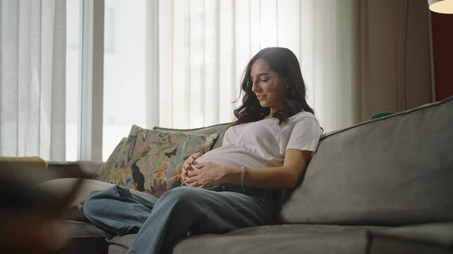A pregnant woman relaxes on a cozy couch in a bright living room, embracing the beauty of motherhood, prioritizing her well-being, and enjoying a peaceful moment during this time of her pregnancy