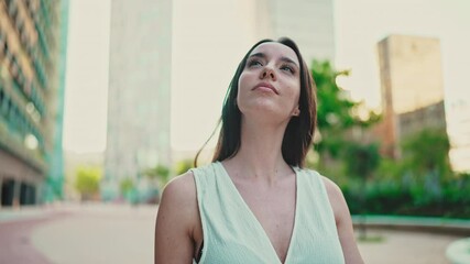 Cute young woman with freckles and dark loose hair wearing white top and jeans is going down the street. Beautiful girl with long eyelashes walks along the street of modern city