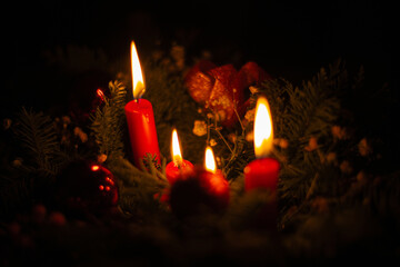 A close-up view of four red candles lit in the dark, surrounded by pine branches, red ornaments, and small flowers, evoking a cozy, festive atmosphere perfect for the holiday season.