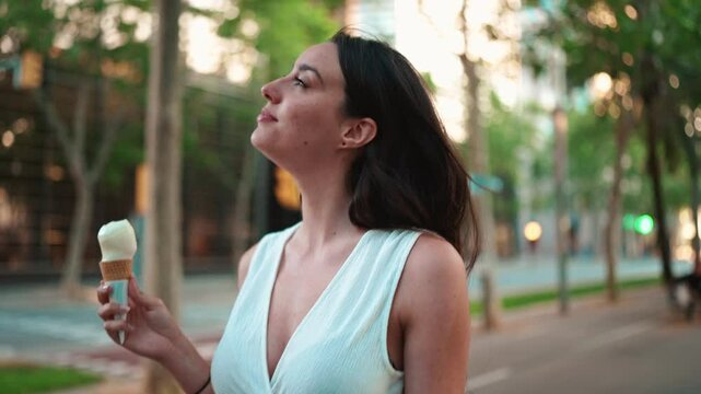 Beautiful woman with freckles and dark loose hair wearing white top is walking down the street and testing ice cream. Cute girl enjoys ice cream on modern city background. Camera is removed