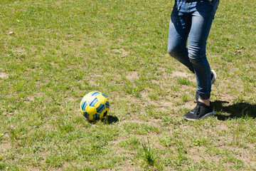 A yellow and blue soccer ball rests on short, slightly patchy grass.  A person in jeans and dark sneakers is walking away, suggesting a game or practice session. The scene is outdoors on a sunny day.