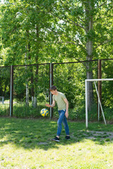 Naklejka premium A young boy playfully juggles a soccer ball outdoors on a grassy field. A soccer goal is visible in the background. Sunlight streams through the trees.