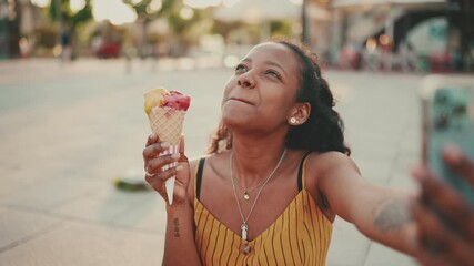 Closeup of smiling young woman with long curly hair with ice cream in her hands making a video call on a mobile phone on an urban city background. Close-up of a happy girl using a smartphone  - Powered by Adobe