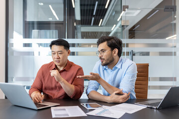 Office workers discussing work project looking at laptop screen, two diverse men sitting at desk...