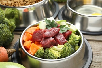 Fresh natural pet food in feeding bowl and vegetables on wooden table, closeup