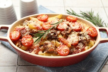 Delicious pasta casserole with cheese, tomatoes, minced meat and rosemary in baking dish on tiled table, closeup