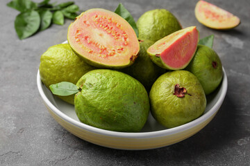 Fresh whole and cut guava fruits in bowl on grey textured table, closeup