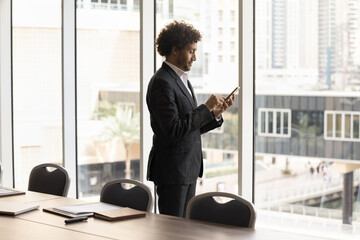 Arabic businessman in formal suit typing message on mobile phone, standing near window in modern...