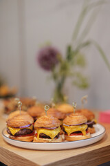 Mini cheeseburgers adorned with melted cheese and fresh toppings rest invitingly on a sunlit wooden table, surrounded by colorful blooms, at a lively event