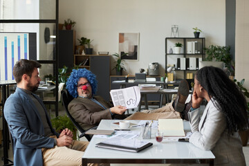 Mature man in formalwear, blue wig and clown nose sitting in front of young employees during discussion of financial papers