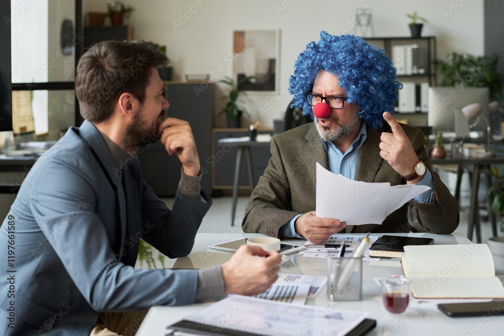 Poster strict mature boss in clown wig and nose looking at laughing male subordinate while explaining him f