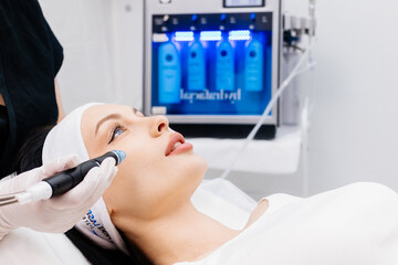 In a cosmetology clinic, a professional female cosmetologist performs a hydrafacial treatment on an...