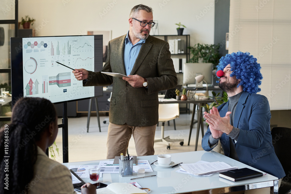 Poster mature male speaker pointing at interactive board and looking at clown businessman clapping hands du