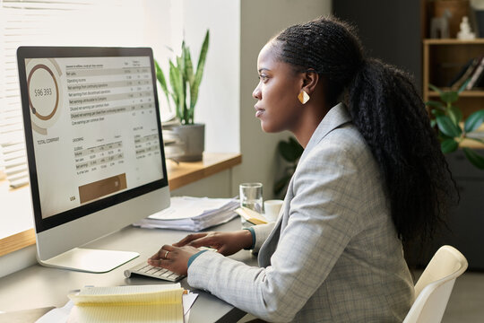 Side view of young serious African American female economist looking at graphic data on computer screen and typing