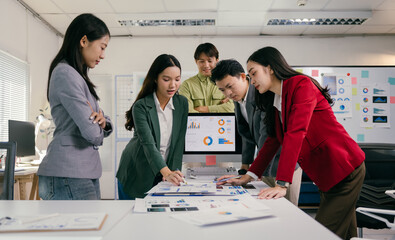 Diverse group of young asian professionals analyzing graphs and reports, collaborating on a project, using computer and printed documents in a modern office environment