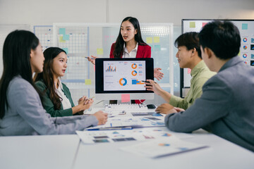 Asian businesspeople are discussing marketing strategy analyzing charts on computer screen during corporate meeting presentation in modern office