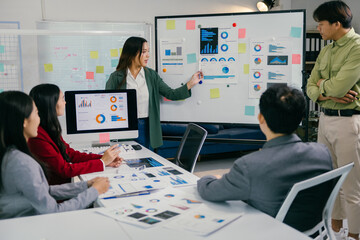Businesswoman presenting marketing strategy on a whiteboard, showcasing charts and statistics while engaging with a team of colleagues during a collaborative office meeting