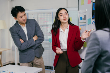 Asian businesswoman in a red blazer leads a meeting. Presenting charts and data on a whiteboard to her colleagues in a modern office setting. Fostering collaboration and strategic decision-making