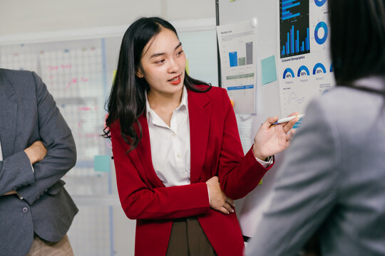 Young asian businesswoman wearing red blazer pointing at charts on whiteboard while leading a meeting with colleagues, discussing marketing data and company strategy
