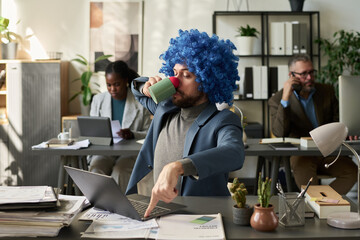 Young white collar worker in clown wig pressing escape key on laptop keypad while drinking coffee...