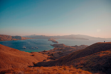Stunning panoramic view of Labuan Bajo, Indonesia, featuring golden rolling hills, a tranquil blue ocean with anchored sailboats, and a vibrant sunset glow. 