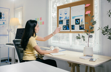 Young professional is pointing at charts with financial data while taking notes at her desk. She is working in a bright modern office