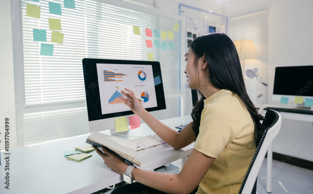 Poster young professional is reviewing data and analytics on her computer while holding a notebook in her o