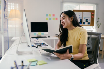 Young professional is smiling while working at her desk, she is holding a notebook and looking at her computer monitor
