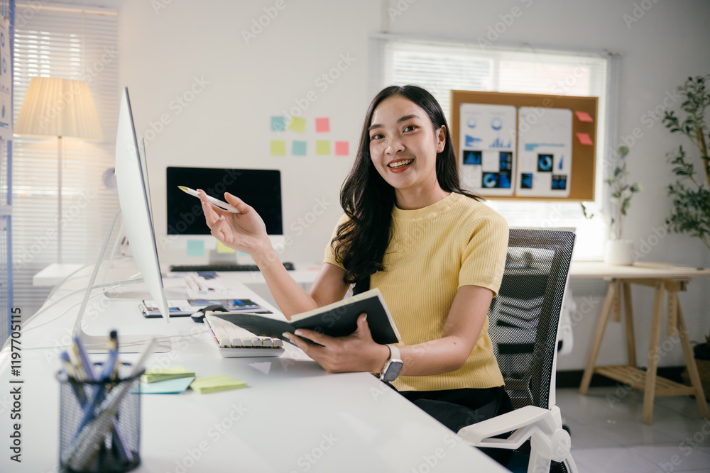 Aufkleber Asian businesswoman is sitting at her desk and holding a notebook while pointing with a pen at a computer screen. She is smiling and appears to be happy with her work
