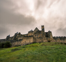 Fototapeta premium Low angle view from a grassy hill of the stone fortress of the medieval castle with stone towers of the medieval fortress of Carcassonne from a hill under a cloudy sky at sunset.
