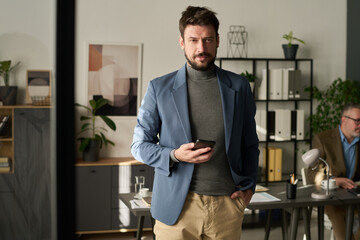 Fototapeta premium Young successful chief executive officer with mobile phone in hand looking at camera against desks of coworkers in office