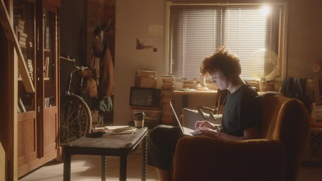 Young man with headphones sitting on couch in cozy, warmly lit apartment with laptop on his lap and typing on keyboard. Side view, zoom shot