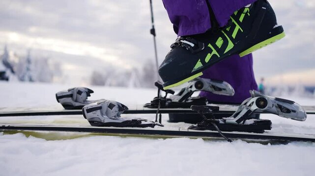 Close-up of skier's legs wearing purple ski pants and black and green ski boots, fastening and unfastening ski bindings on snowy mountain top