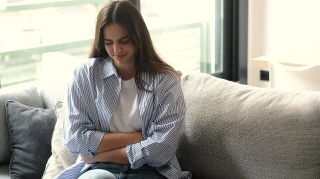 Young brunette woman sitting on a couch holding her stomach tightly in pain, showing symptoms of severe menstrual cramps, digestive discomfort, endometriosis, or abdominal tension caused by colic