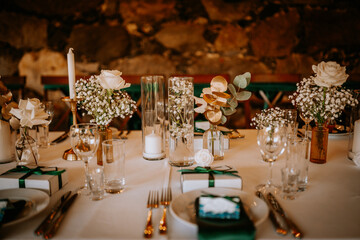 Elegant wedding table with white roses, candles, greenery, and gift boxes on a white tablecloth, creating a romantic and refined atmosphere.