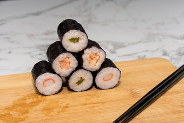 Stack of sushi rolls with various fillings, including tuna, shrimp and cucumber. The rolls are stacked one on top of the other to form a pyramid and sits on wooden cutting board with chopsticks nearby