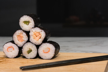 Stack of sushi rolls with various fillings, including tuna, shrimp and cucumber. The rolls are stacked one on top of the other to form a pyramid and sits on wooden cutting board with chopsticks nearby