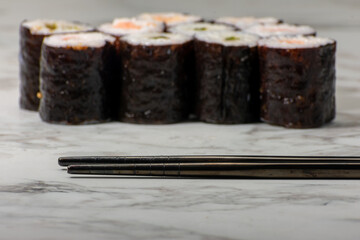 Chopsticks in the foreground with sushi rolls in the background, all placed on a clean white surface. The minimalistic setting highlights the elegance and simplicity of Japanese cuisine.