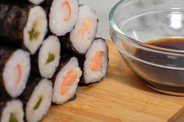 Glass bowl filled with soy sauce sits beside a neatly arranged pyramid of sushi rolls, each featuring a different type of fish. Elements rest on wooden board, creating a warm and inviting composition