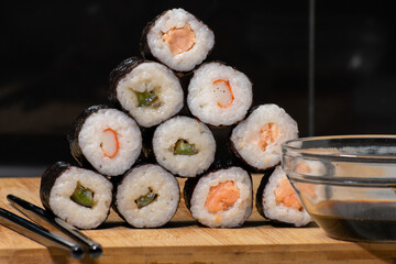 Glass bowl filled with soy sauce sits beside a neatly arranged pyramid of sushi rolls, each featuring a different type of fish. Elements rest on wooden board, creating a warm and inviting composition