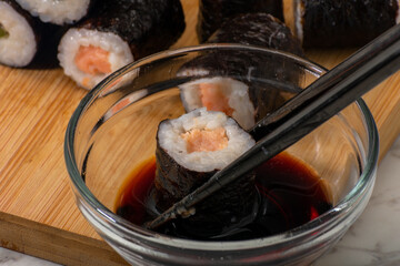 Chopsticks grasp a piece of sushi dipped in soy sauce within a clear glass bowl. In the background, various sushi rolls are arranged on a wooden surface, showcasing Japanese culinary artistry.