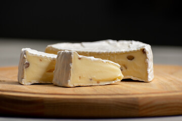 Wheel of creamy camembert cheese topped with nuts, resting on a rustic wooden surface against a solid black background. The rich textures and contrast create an elegant culinary composition