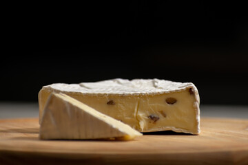 Wheel of creamy camembert cheese topped with nuts, resting on a rustic wooden surface against a solid black background. The rich textures and contrast create an elegant culinary composition