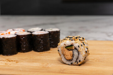 Sushi roll sits on a wooden cutting board. The sushi roll is surrounded by other sushi rolls, creating a sense of abundance and variety