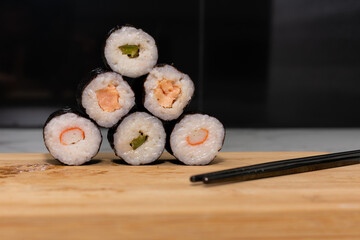 Stack of sushi rolls with various fillings, including tuna, shrimp and cucumber. The rolls are stacked one on top of the other to form a pyramid and sits on wooden cutting board with chopsticks nearby