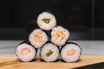Stack of sushi rolls with various fillings, including tuna, shrimp and cucumber. The rolls are stacked one on top of the other to form a pyramid and sits on wooden cutting board with chopsticks nearby