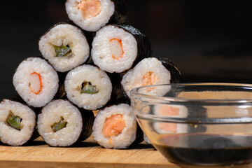 Glass bowl filled with soy sauce sits beside a neatly arranged pyramid of sushi rolls, each featuring a different type of fish. Elements rest on wooden board, creating a warm and inviting composition