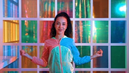 Happy hispanic girl in lively mood moving along music in neon light while wearing colorful cloth at night club. Street dancer making energetic footstep or movement while looking at camera. Regalement.